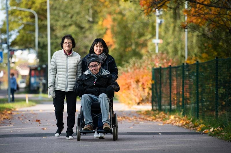 Kund i rullstol på promenad tillsammans med sin fru och en medarbetare från hemtjänsten
