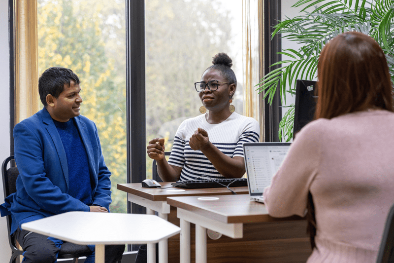 Three colleagues talking in an office, one gesturing while seated at a desk with a computer.