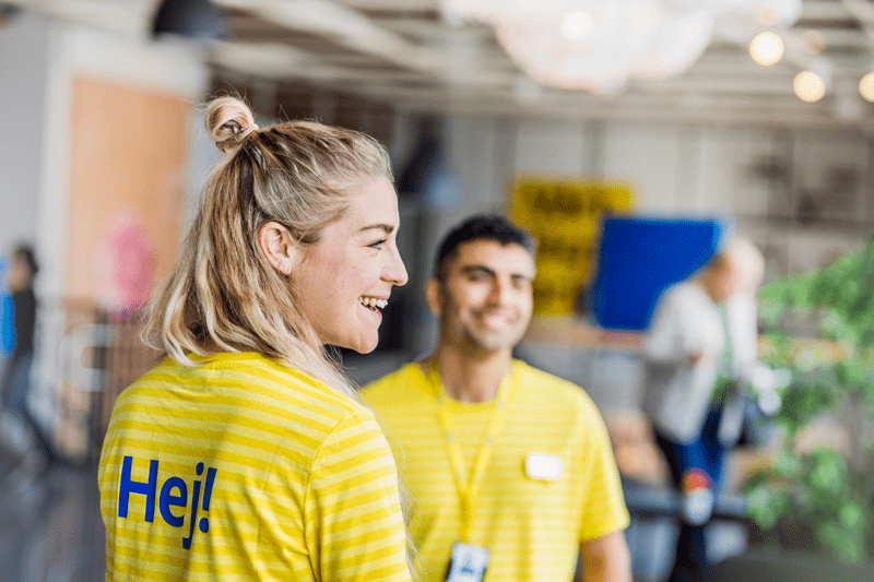 Smaidošs IKEA darbinieks dzeltenā uniformā ar uzrakstu „Hej” uz muguras, fonā redzams vēl viens darbinieks.