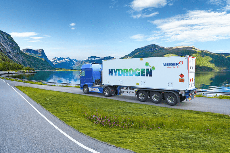 Truck with a trailer labeled 'HYDROGEN' and 'MESSER Gas for Life' driving on a road beside a lake with mountains in the background