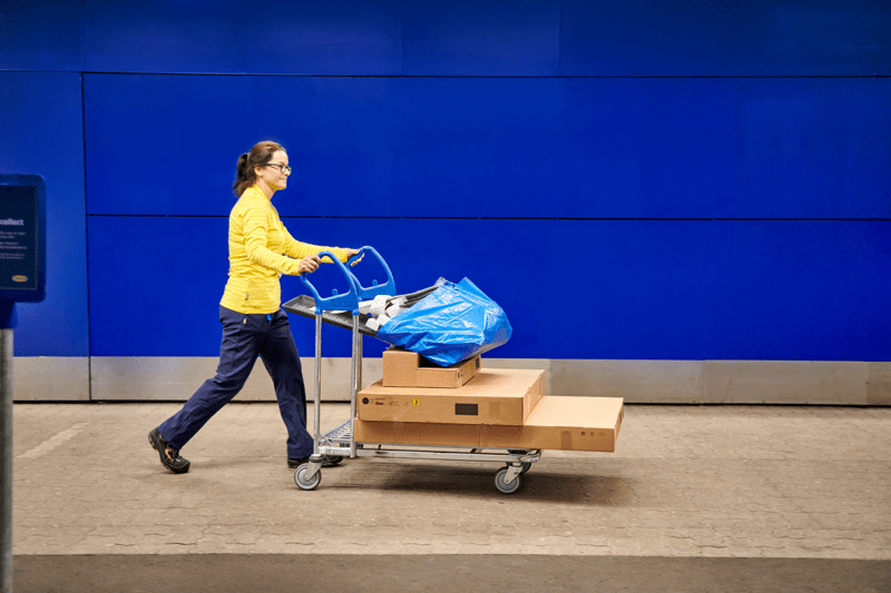 IKEA darbuotoja eina prie parduotuvės sienos ir stumia vežimėlį su supakuotomis prekėmis, iliustruodama e. užsakymų atsiėmimo paslaugą.