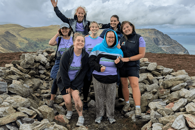 A group of people wearing matching purple 'Walks for Women' T-shirts pose beside a stone wall on a mountain summit, with rolling hills and the sea visible in the background. Text overlay reads “Our Impact”.
