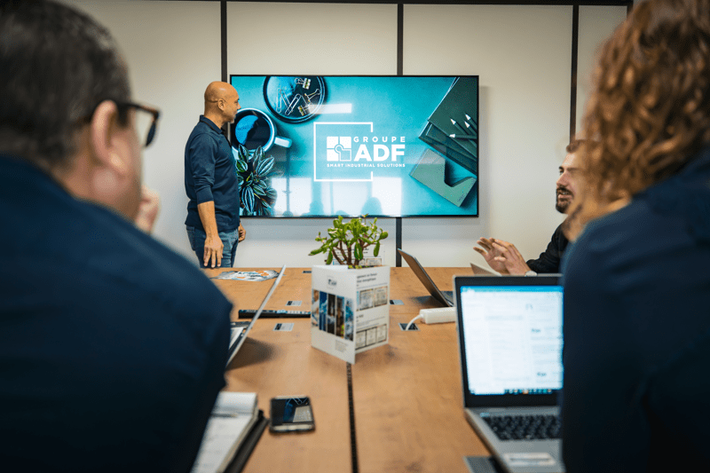 Un intervenant anime une réunion de travail face à des collaborateurs autour d'une table, devant un grand écran affichant le logo du Groupe ADF
