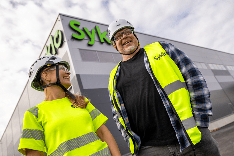 Kaksi työntekijää seisoo Syklon rakennuksen edessä, käyttäen turvaliivejä ja kypäriä.