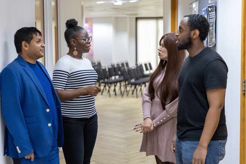 Four colleagues standing in a hallway having a conversation, with rows of chairs visible in the background