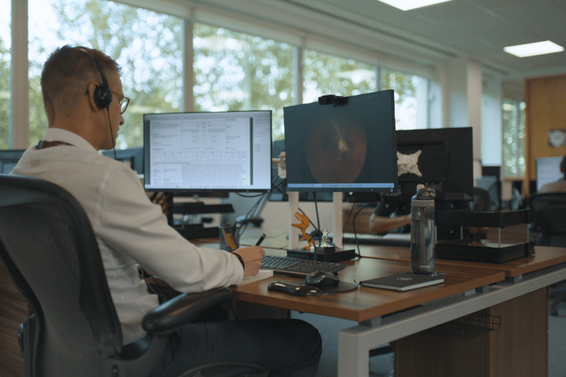 Man sitting at desk with two screens, one screen has a fundus camera image on it. Background is of people sat at their desks working 