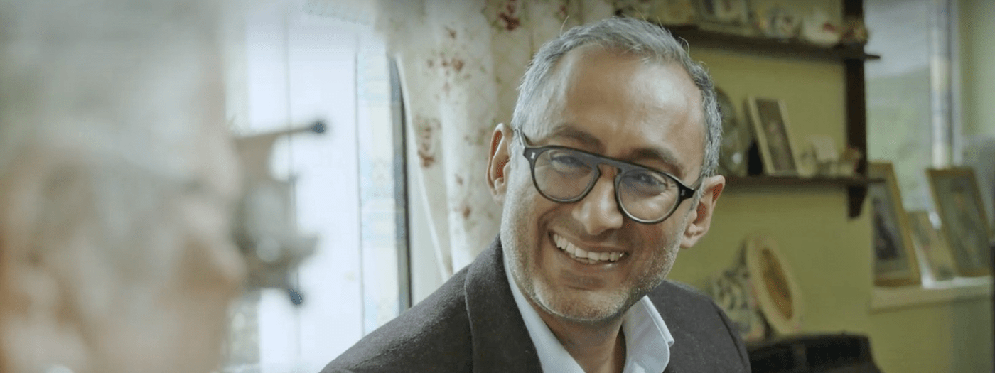 Mam, smartly dressed with glasses on smiling, he is talking to someone and behind him are curtains and shelves with photos on