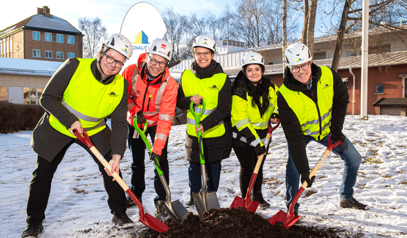 Fem personer med hjälmar och skyddsvästar håller i varsin spade mot en jordhög en solig vinterdag.