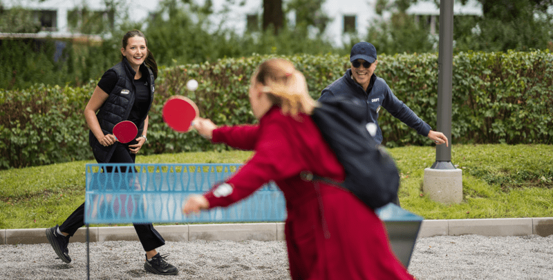 Tre personer som spelar pingis utomhus med grönska i bakgrunden.