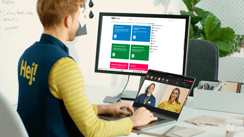 IKEA darbuotojas sėdi darbo vietoje prie nešiojamojo kompiuterio su papildomu monitoriumi ir dalyvauja vaizdo skambutyje; ant jo uniformos nugaros matosi užrašas „Hej!“