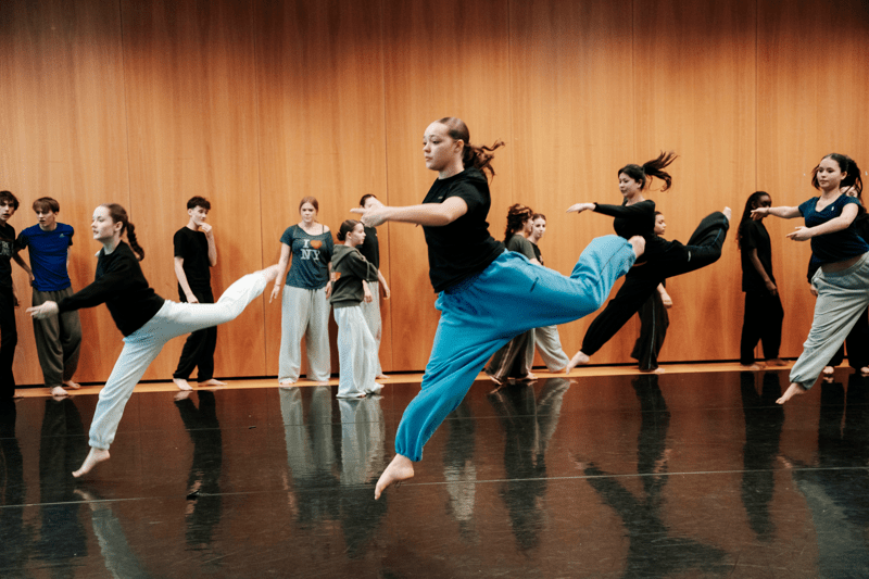Four dancers practising jumps in a studio while others watch them.