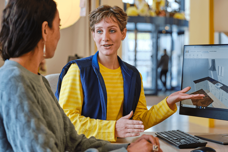 IKEA darbuotoja su dryžuota geltonos spalvos uniforma ir mėlyna liemene pasakoja pirkėjai apie skaitmeninį virtuvės planą kompiuterio ekrane.