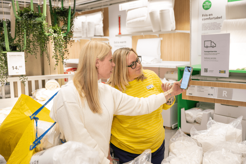 Geltonos spalvos uniformą vilkinti IKEA darbuotoja padeda pirkėjai nuskaityti QR kodą nuo plakato parduotuvėje.