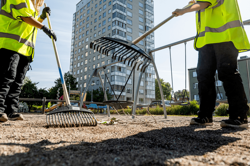 Två personer med varselvästar krattar i en sandlåda, med ett bostadshus i bakgrunden.