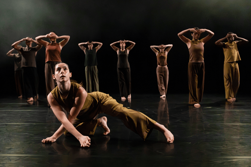 A group of contemporary dancers perform on a dark stage. One dancer in the foreground crouches low with one leg extended and looks upward, while several others stand behind in a line with their arms raised and hands covering their eyes, all wearing earth-toned outfits under soft stage lighting.