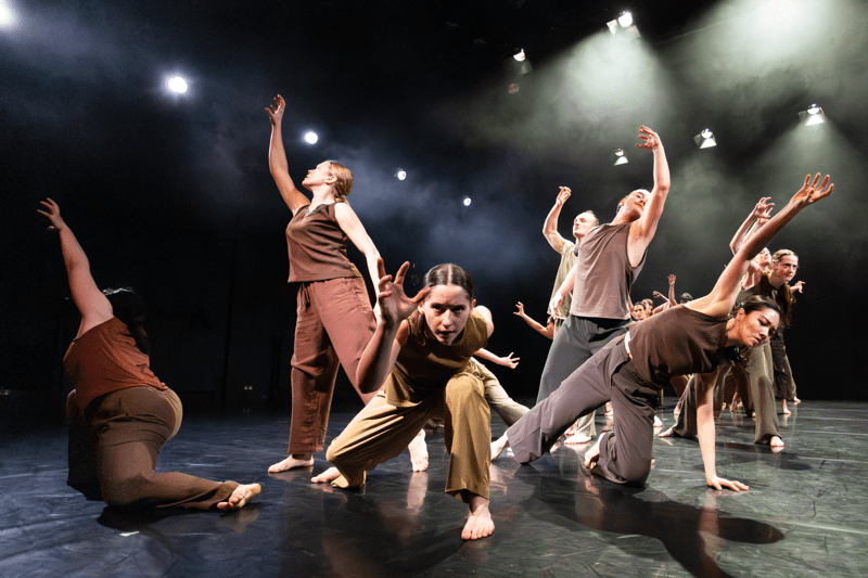 A group of contemporary dancers perform energetically on a stage under bright overhead lights. In the foreground, one dancer crouches low and reaches forward with an intense expression, while others around them extend their arms upward and outward in expressive poses. All are barefoot and dressed in coordinated earth-toned outfits, with a dark, smoky stage background enhancing the dramatic lighting.