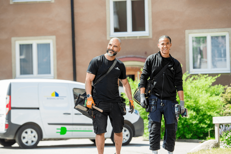 Två män i arbetskläder går framför ett bostadshus.