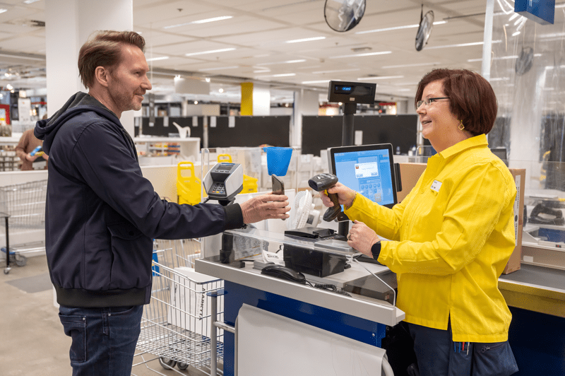 Naeratav kollases särgis IKEA töötaja teenindab külastajat, skannides kassas tooteid.