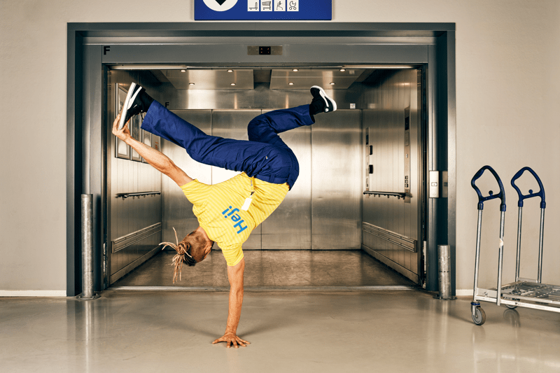 Kollases särgis ja tumesinistes pükstes IKEA töötaja hoiab ühe käe peal tasakaalu, tema taustaks on avatud ustega lift, mille kõrval on kaks ostukäru.