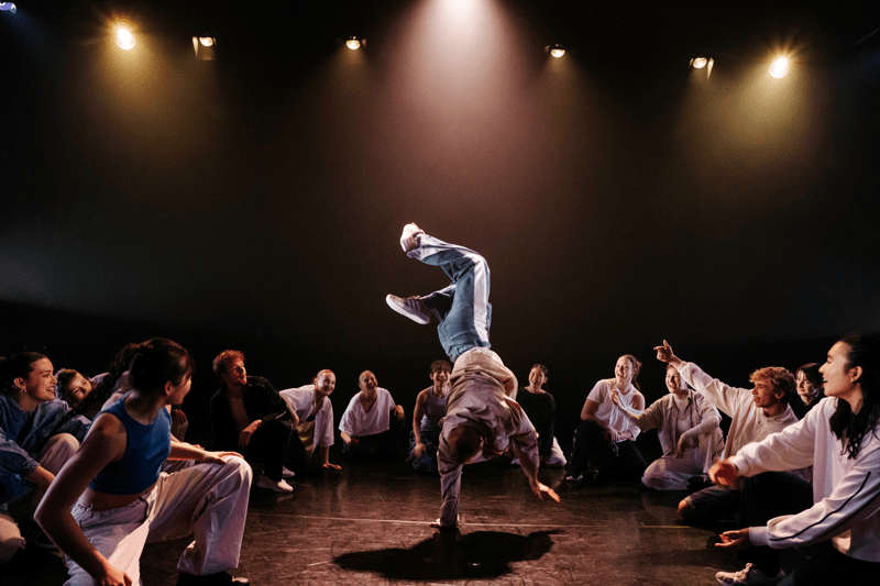 A dancer performs a dynamic breakdance handstand at the centre of a circle of spectators on a dark stage. Under warm spotlights, the surrounding group watches, smiles, and gestures enthusiastically, creating an atmosphere of energy, encouragement, and shared participation.