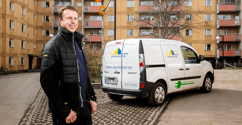 En man står framför ett gult flerbostadshus och ler mot kameran, bredvid en bil med Signalistens logga.