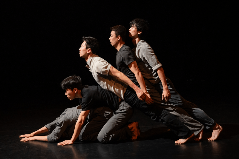 Five dancers on a black stage facing left. They are barefoot and wearing shades of grey and black. They are leaning left stacked on top of each other.