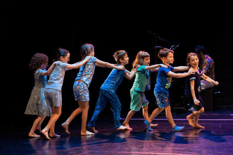 Seven children wearing blue clothing on a dance stage in a line facing right with their hands on the person in front.