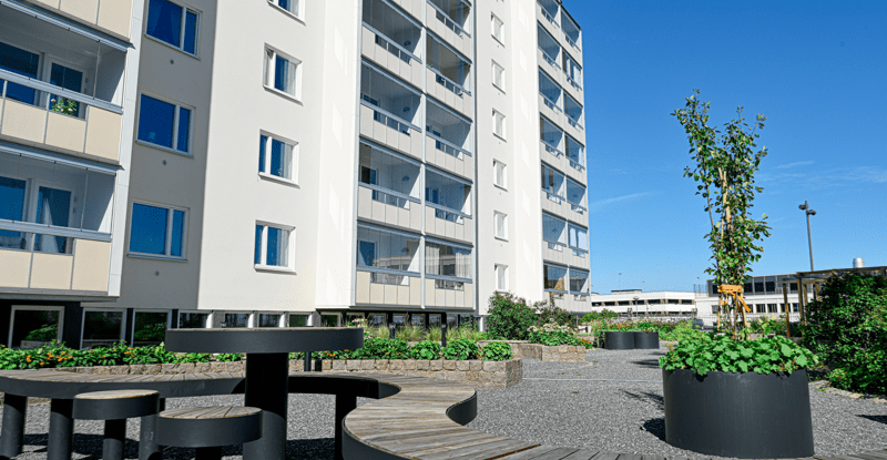 Ljust flerbostadshus och en innergård med sittplatser och grönska.