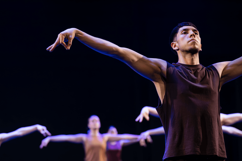 A male dancer in a sleeveless dark top performs on stage with his arms extended in a graceful pose, lit by blue and purple stage lighting. Several other dancers appear blurred in the background, echoing similar arm movements against a dark backdrop.