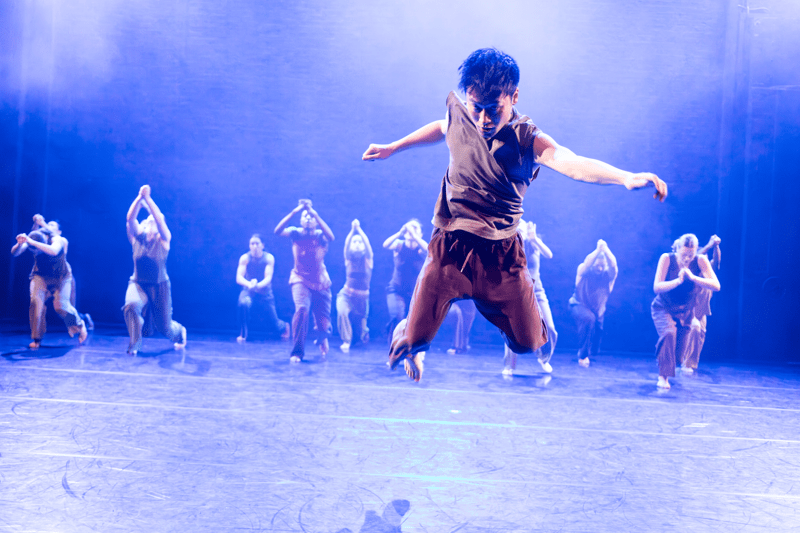 A dancer leaps powerfully across a stage under bright blue-white lighting, arms outstretched and knees bent mid-air. Behind him, a group of dancers kneel in formation with their hands raised together, partially blurred against the dimly lit theatre backdrop.