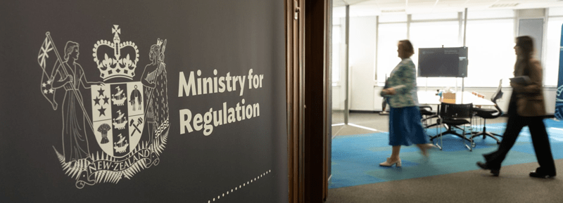 Close up of a Ministry for Regulation logo on a banner. In the background there are blurred people walking past in an office environment. 